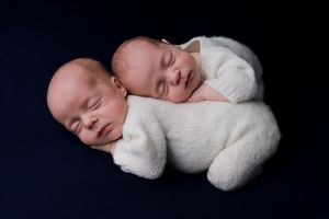 Twin babies in white outfits sleeping contentedly against a dark backdrop.