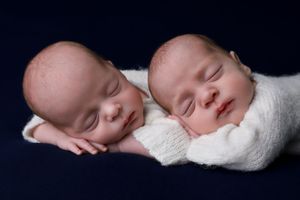 Sleeping twins in white sweaters resting head-to-head on a dark background.