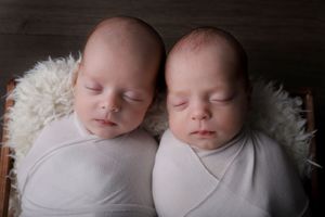 Twin newborns swaddled in white wraps sleeping side by side on furry white bedding.
