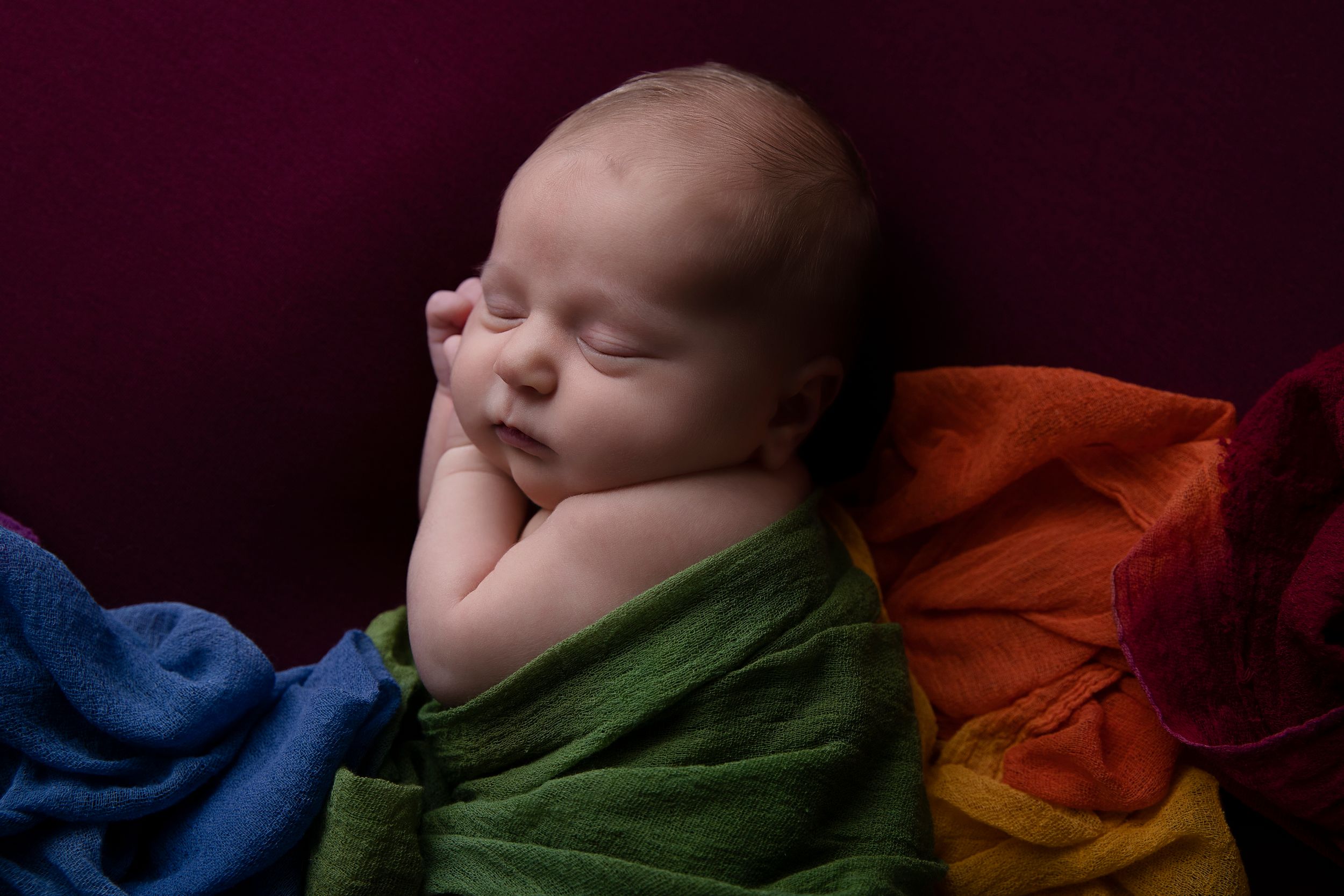 Sleeping newborn baby on colorful rainbow fabric against dark background.