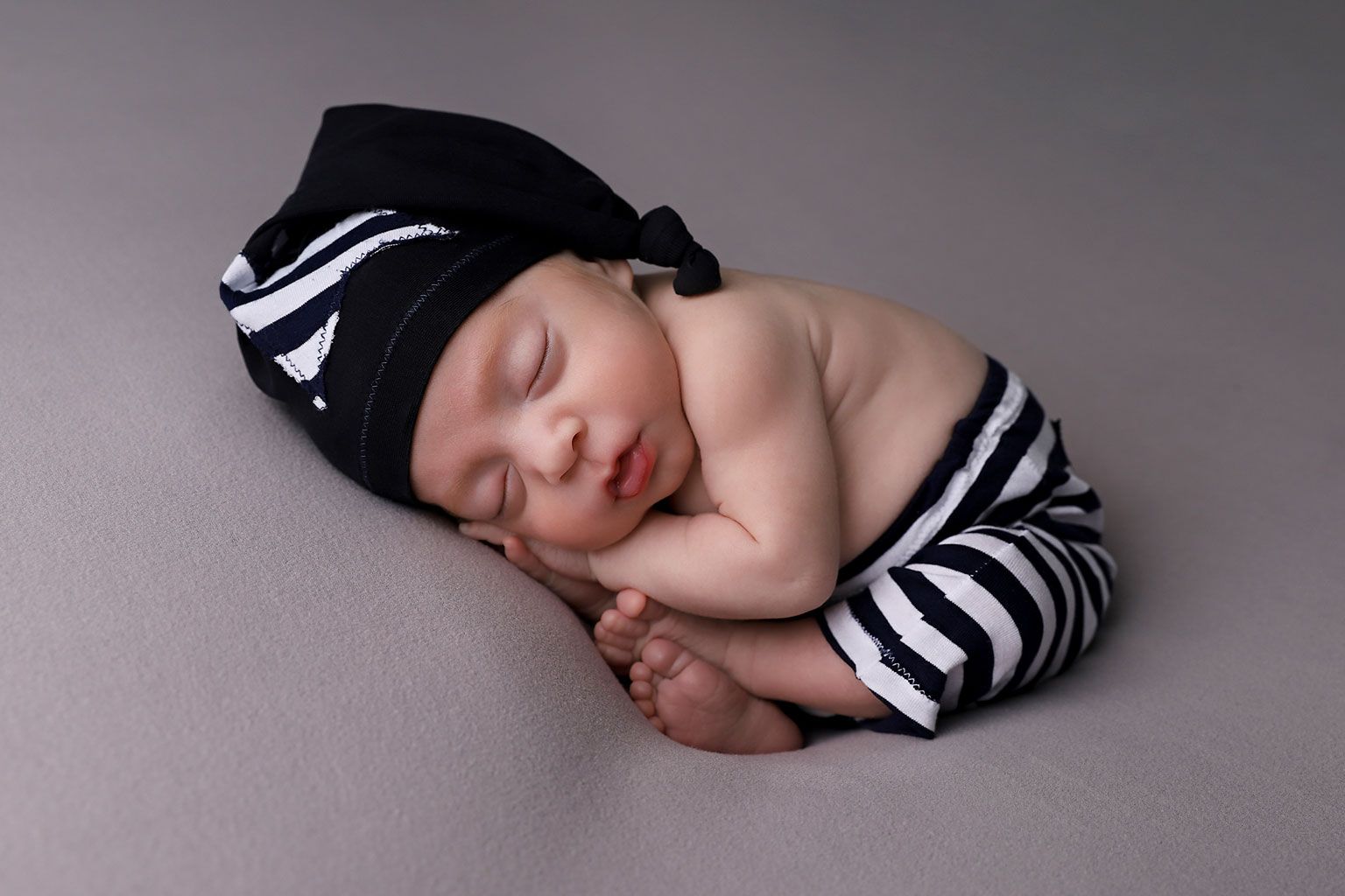 Sleeping newborn wearing striped legwarmers and black beanie on gray background.