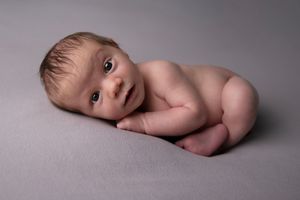 Newborn lying on side on gray backdrop looking at camera with wide eyes.