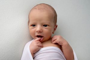 Newborn baby in white swaddle with curious expression sucking on fingers.