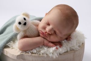Nouveau-né endormi, blotti avec une petite peluche ours sur une douce couverture blanche dans un contenant en bois beige.