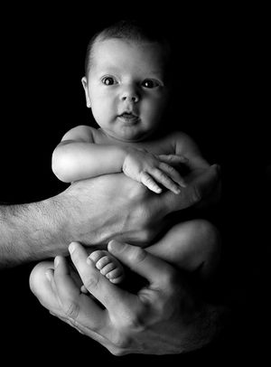 Portrait artistique en noir et blanc d’un nourrisson soutenu par des mains bienveillantes du papa sur un fond sombre.