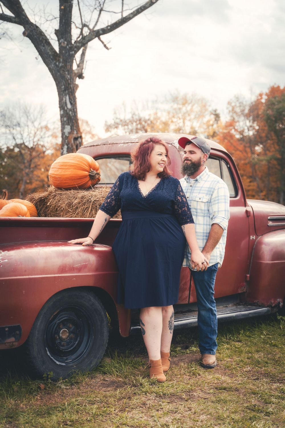 Couple poses with vintage red pickup truck in rustic outdoor setting with pumpkin decoration.