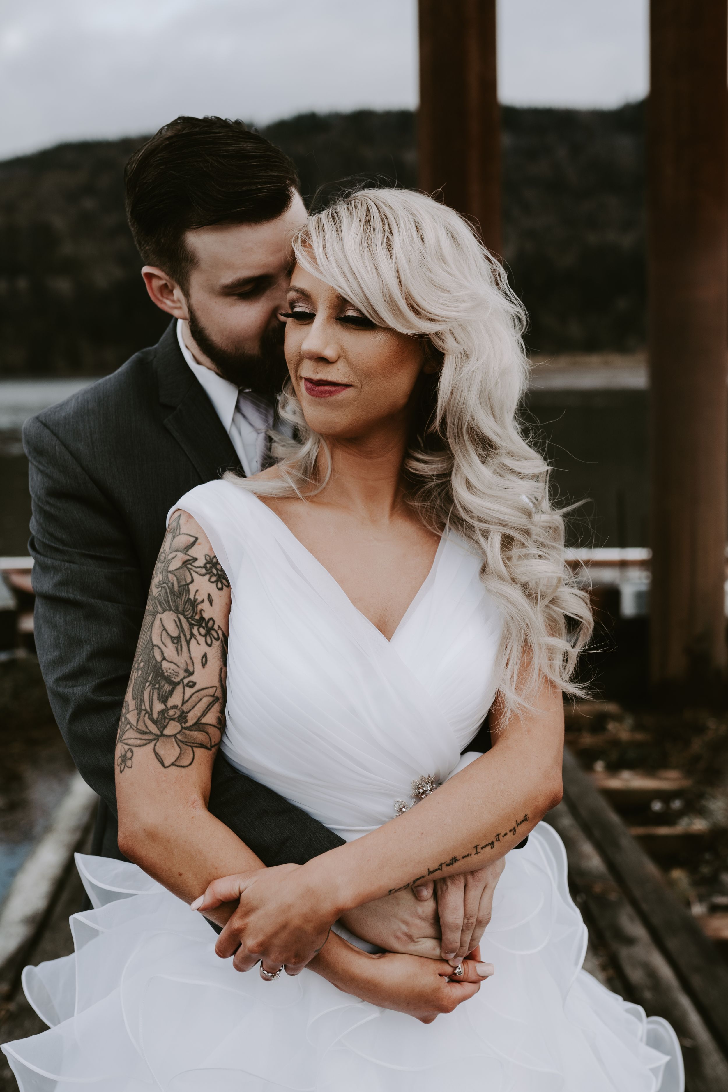 Intimate wedding portrait showing bride in white dress with visible tattoos embracing groom against moody backdrop.