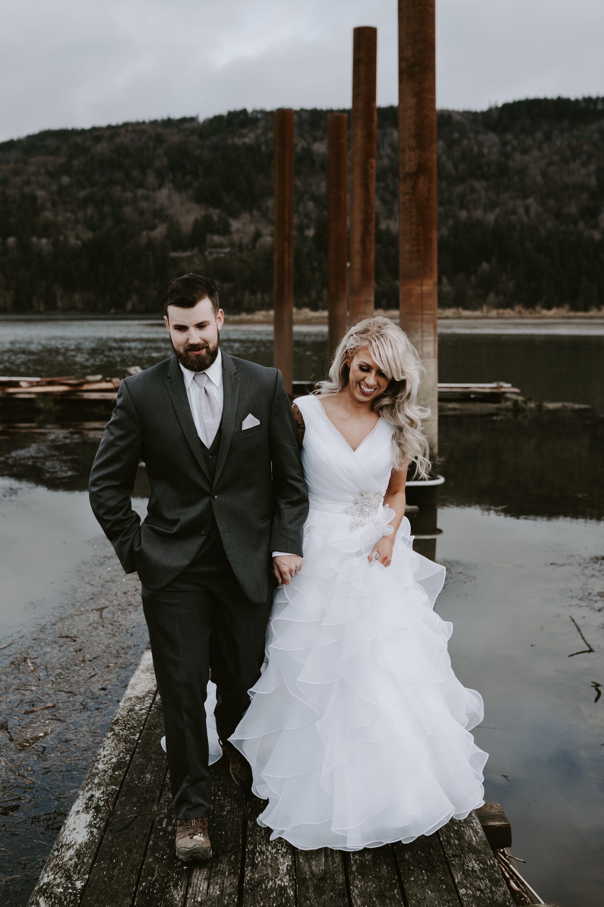 Couple in wedding attire posing on wooden dock with mountain lake and rustic poles in background.