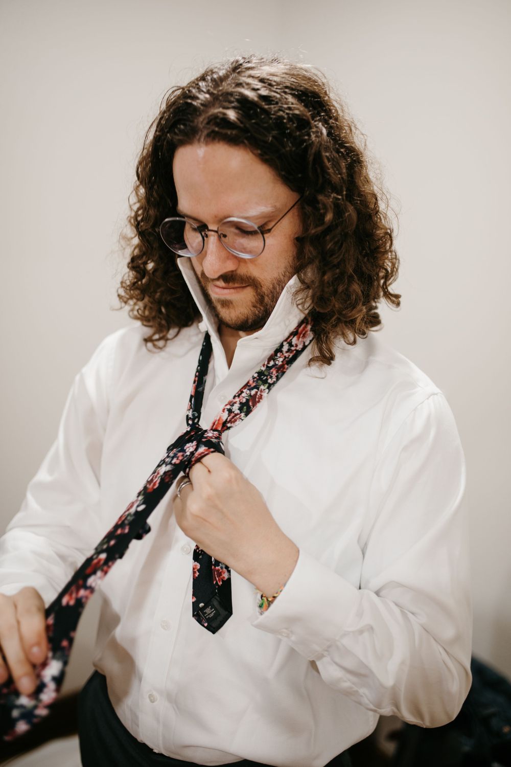 Man with curly hair and glasses adjusts his floral tie while wearing a white dress shirt.