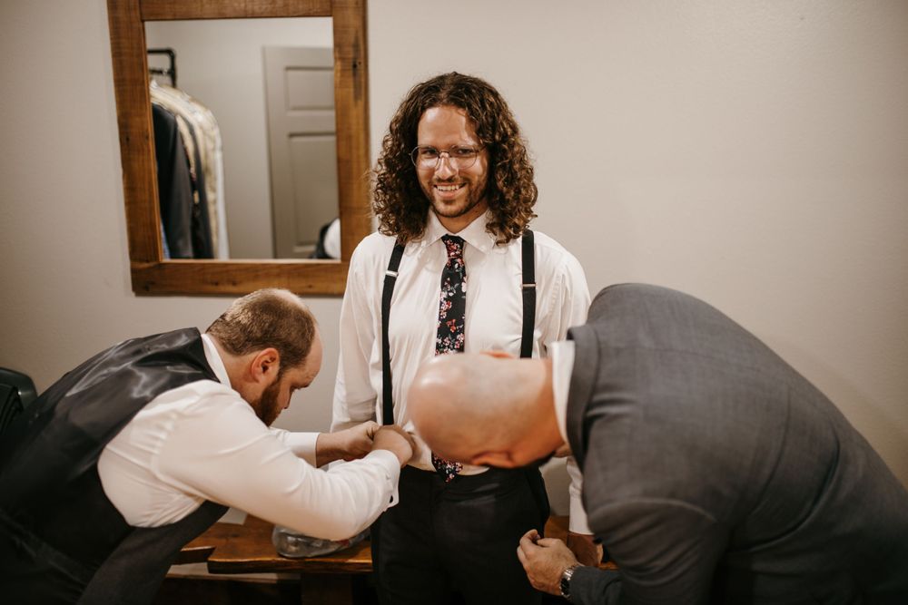 Groom with curly hair smiles as groomsmen help him get dressed in suspenders and floral tie.