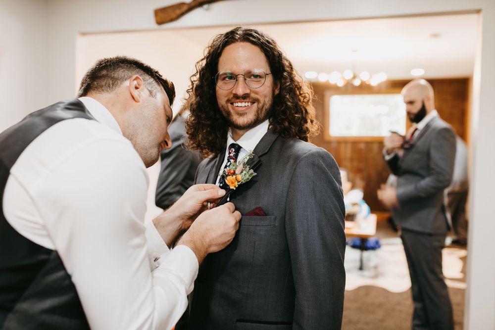 Groomsman pins boutonniere on smiling groom in gray suit with curly hair and glasses.