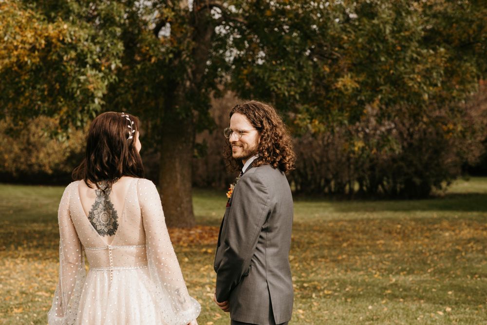 Groom in gray suit smiles at bride in elegant lace cape gown during outdoor first look.