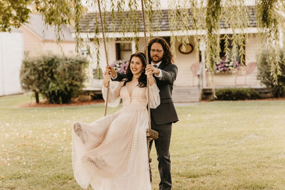 Groom pushes bride on garden swing in blush tulle gown outside historic farmhouse.