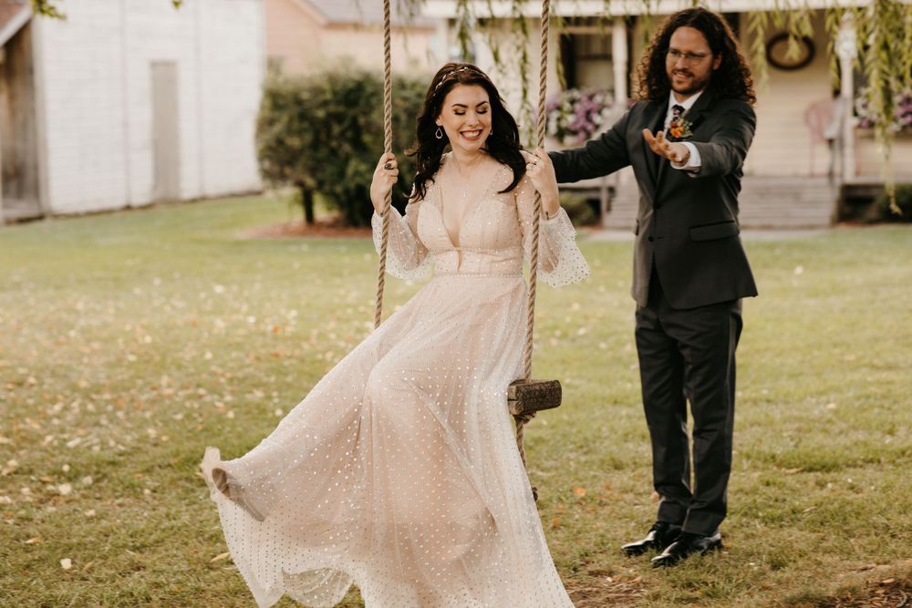 Bride laughs on rustic swing in blush gown as groom pushes her on lush green lawn.