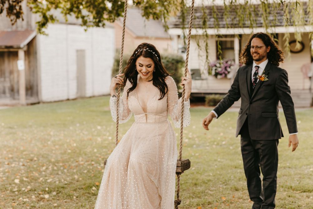 Bride in blush sparkle gown enjoys garden swing as groom walks alongside her smiling.