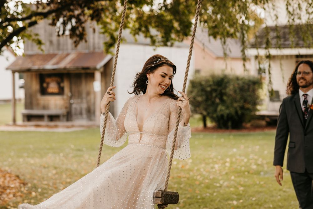 Bride in lace wedding gown smiling on a rope swing at rustic outdoor fall wedding venue.