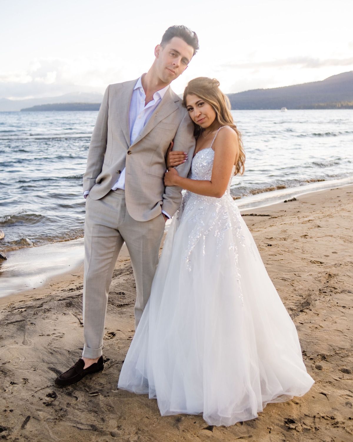 A couple in formal wedding attire poses together on a beach at North Lake Tahoe Event Center with mountains in the background.
