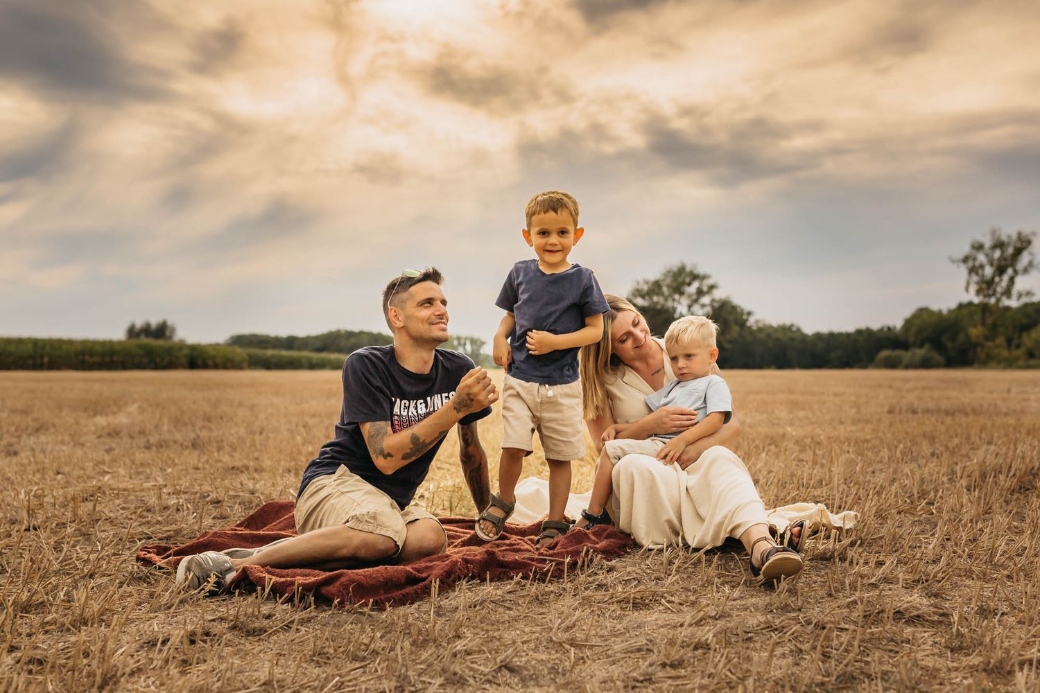 Family sitting on blanket in golden wheat field during sunset for outdoor portrait session.