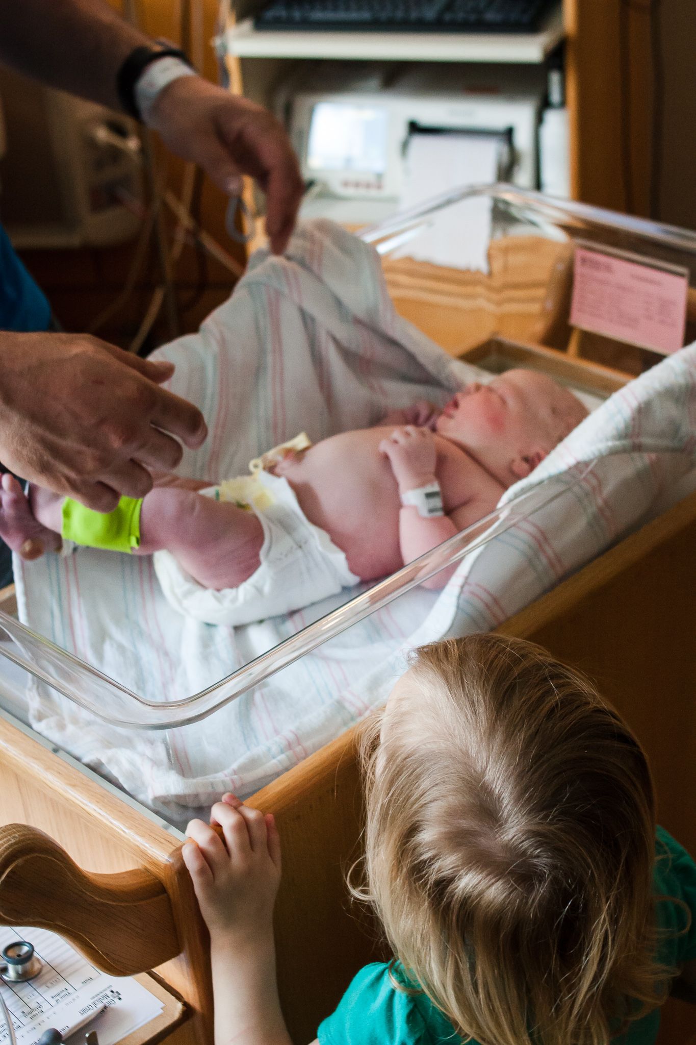 Big sister taking a peek at her new baby sister while dad changes the baby's diaper in the hospital bassinet.