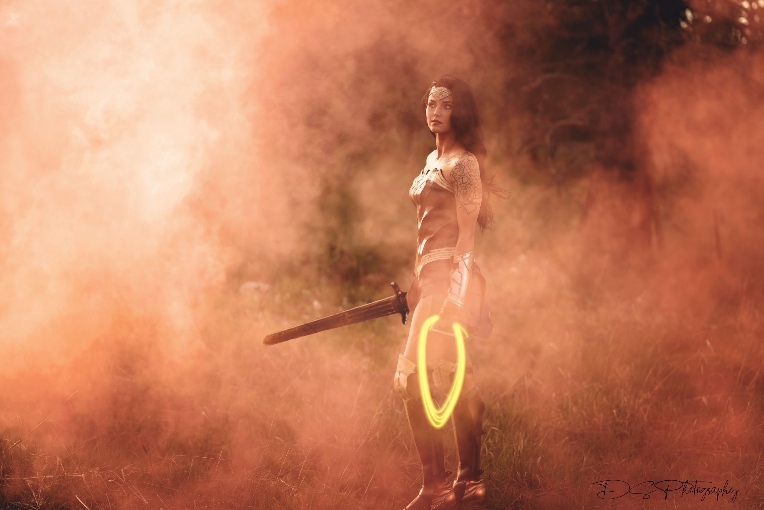 Figure in athletic wear stands dramatically amidst orange smoke holding illuminated rings at sunset.