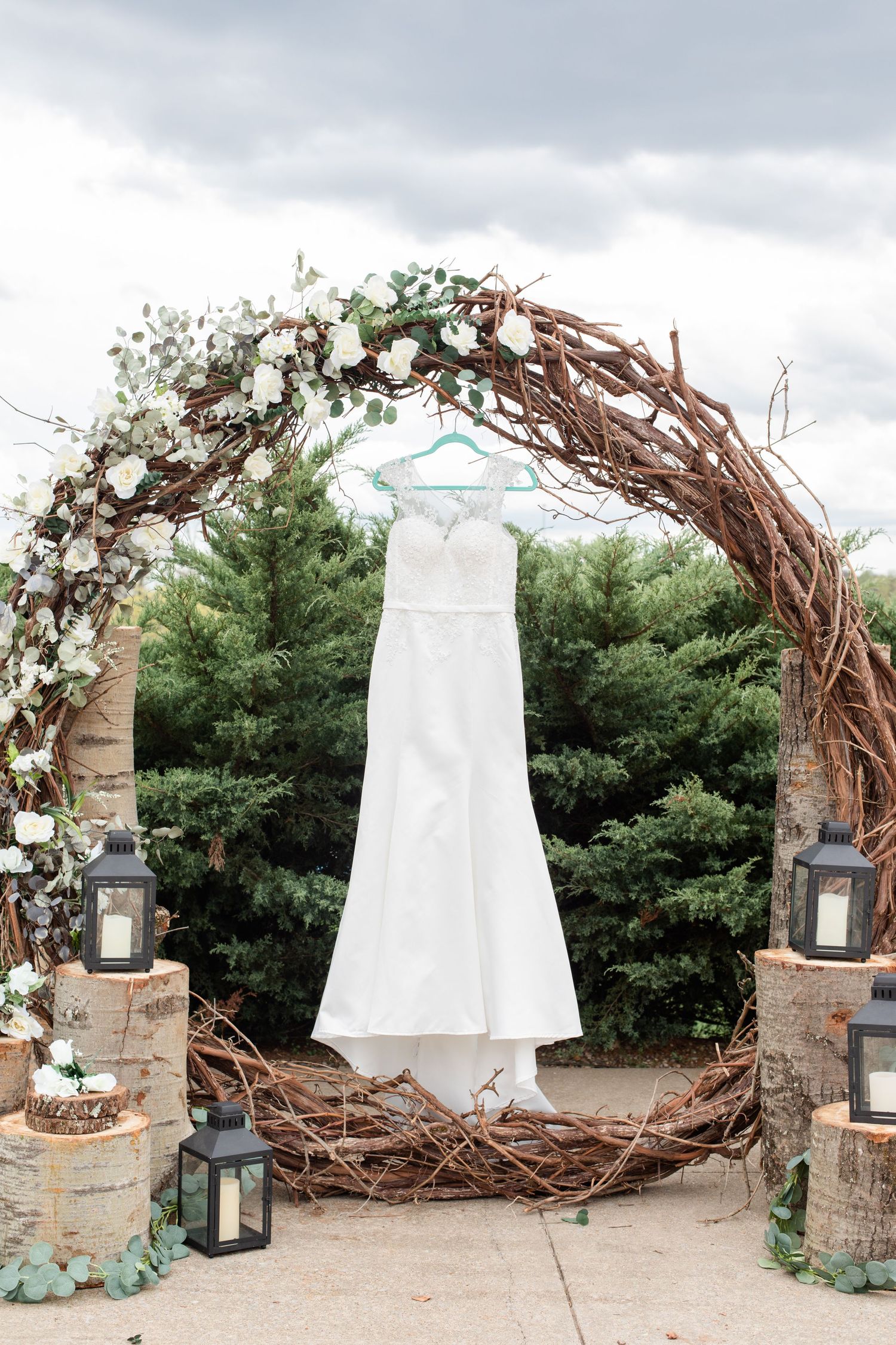 A white wedding dress hangs from a rustic wooden arch decorated with lanterns and flowers.