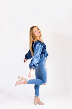 Woman in blue satin top and jeans poses playfully against white background.