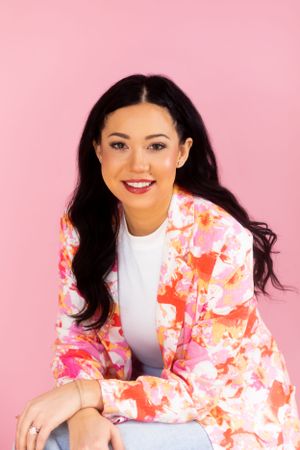 A woman in a coral floral blazer and white top smiles against a pink background.