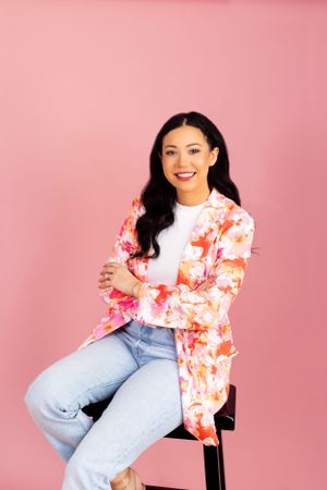 A woman in a floral pink blazer and light denim jeans poses on a stool against a pink background.