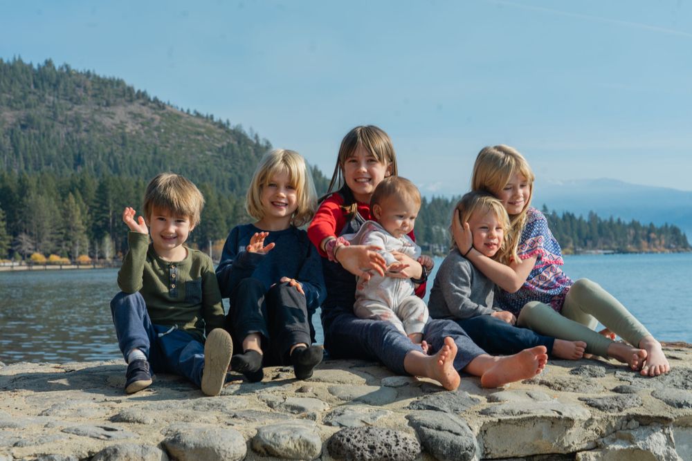 Family group sits together on rocky shore with lake and mountains in background.