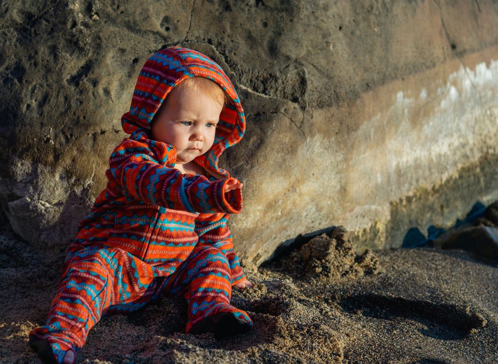 A small figure in striped orange winter wear sits on rocky beach during golden hour.