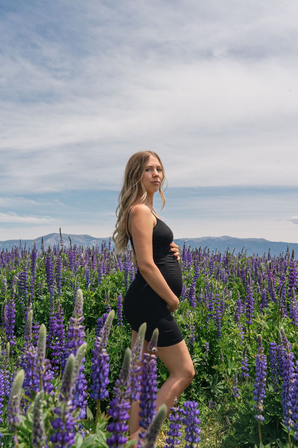 Person in black dress poses among blooming purple lupines with mountains in the background.