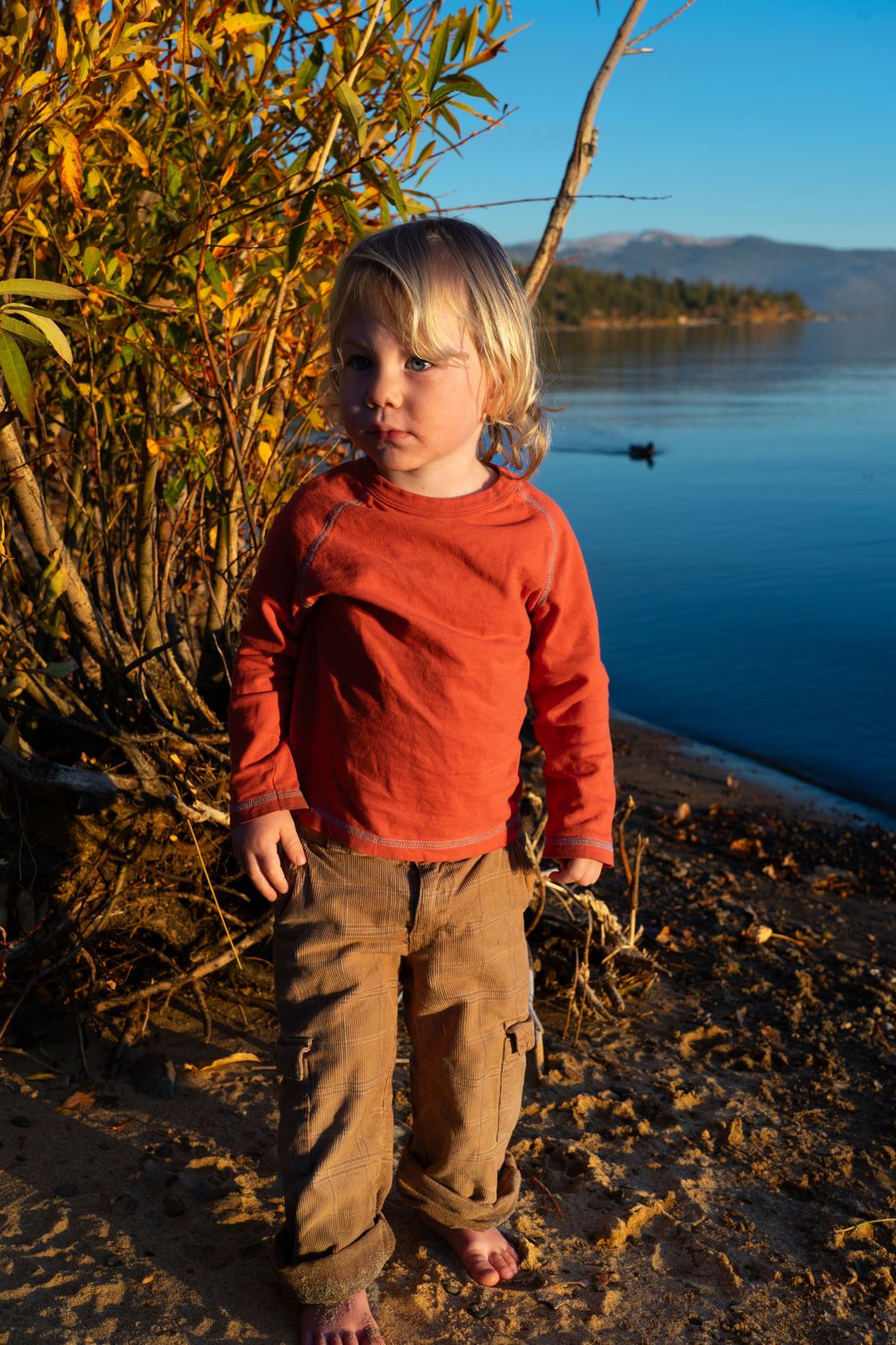 A small figure in red shirt and khaki pants stands by a serene lake with mountains in the background.