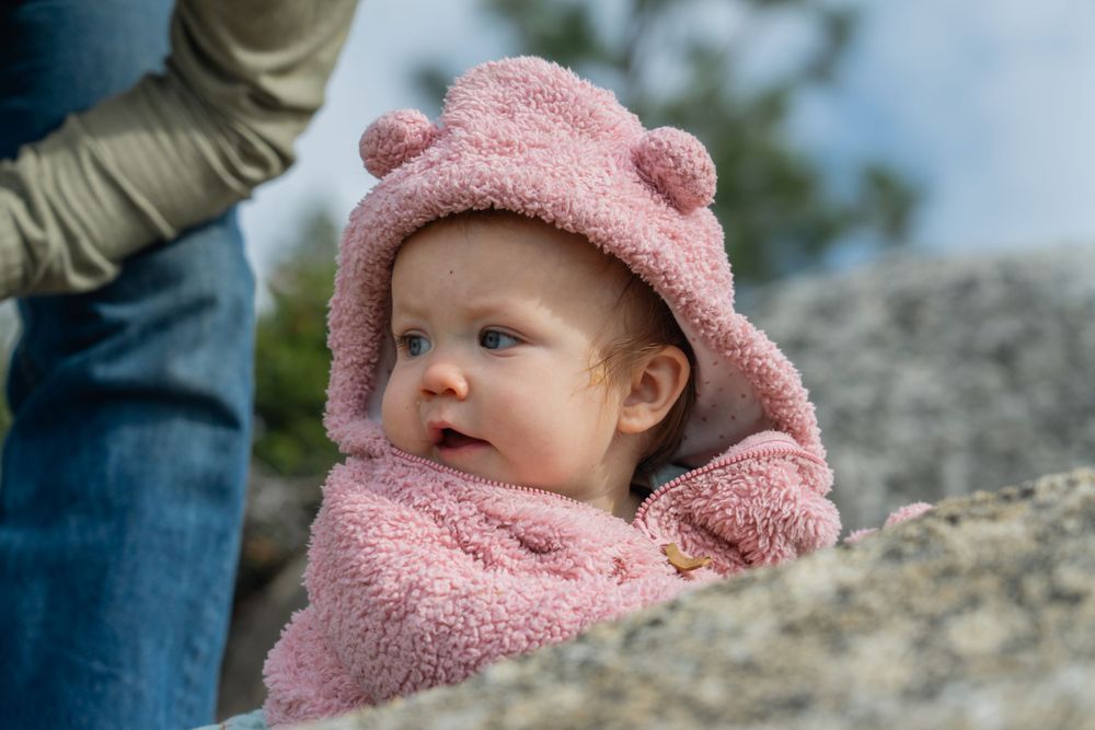 Outdoor natural light Baby wearing a cozy pink fleece hat and jacket sits outdoors against a rocky background.