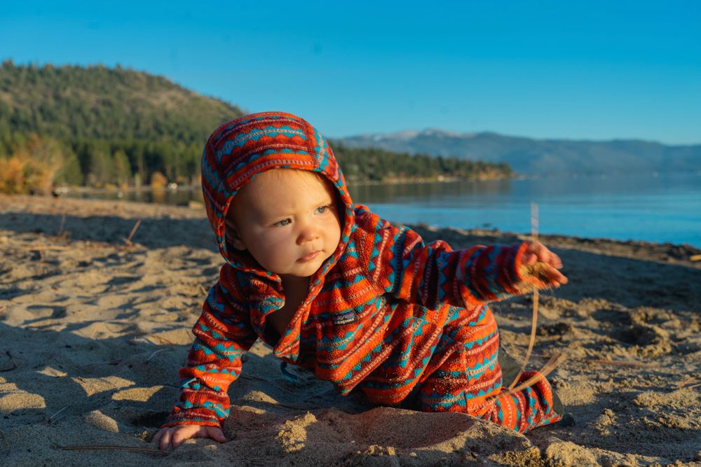 A small explorer in orange patterned coat plays on a beach with mountains in the distance.