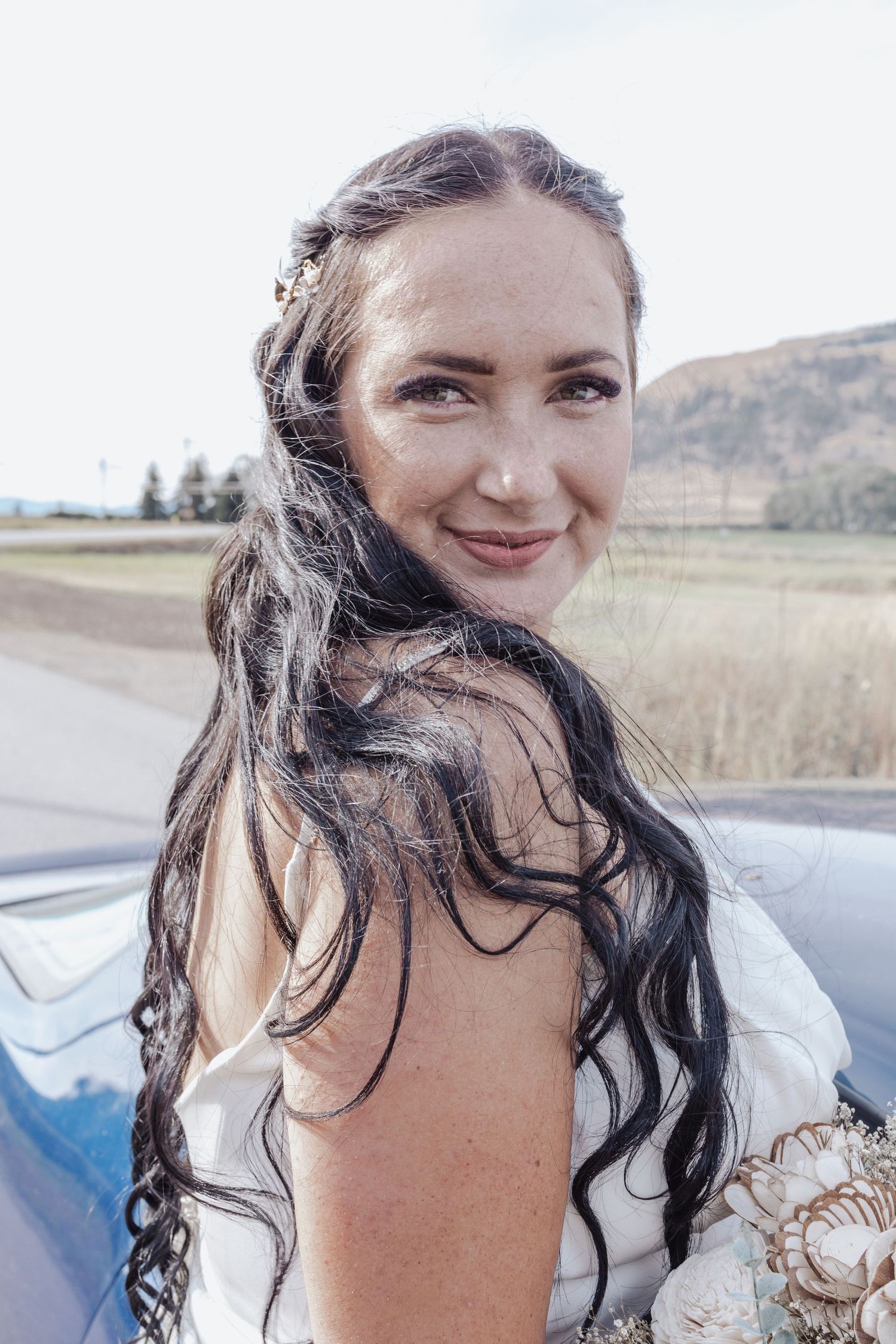 Bride with flowing dark hair smiling over shoulder in countryside setting.