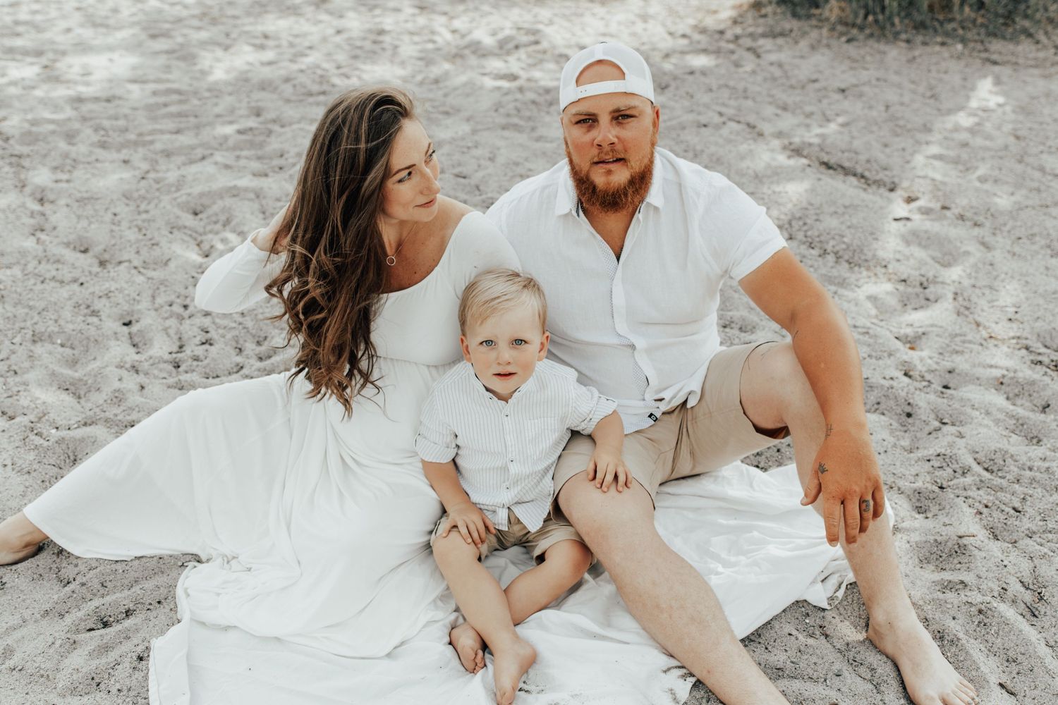 Family dressed in white sits together on sandy beach during sunset photo session.