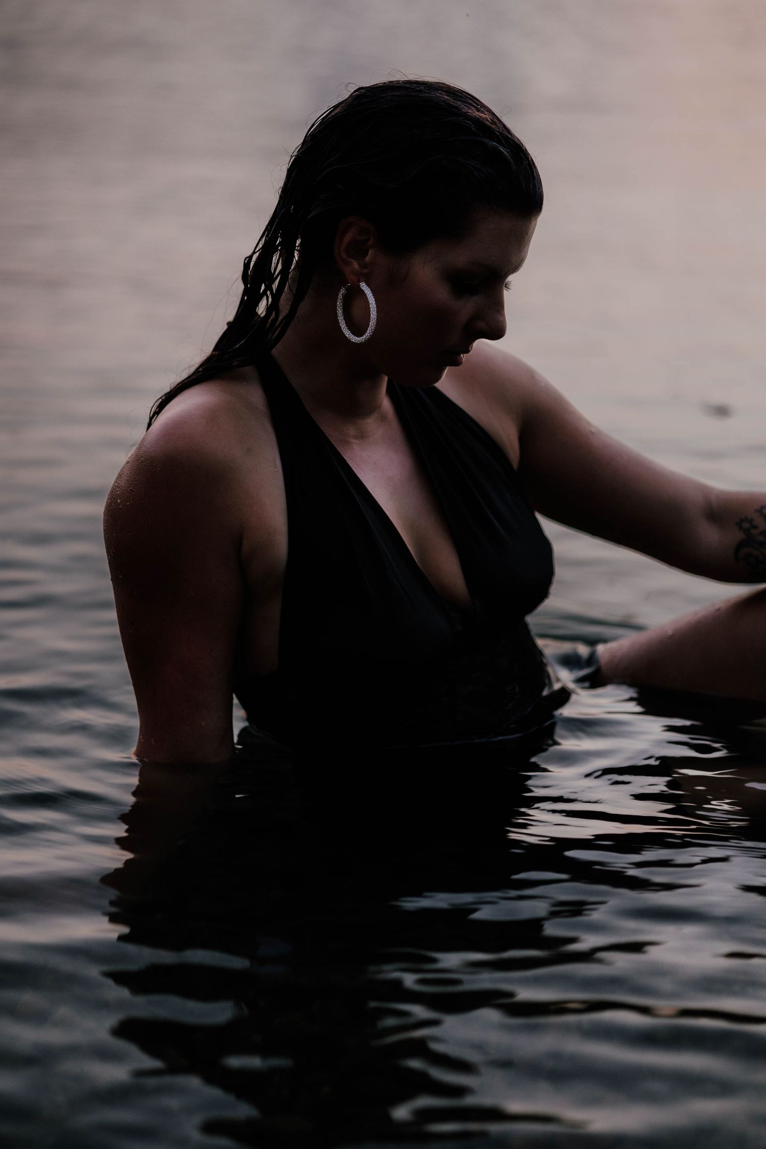 Person wearing black swimsuit and hoop earrings wading in calm water at sunset.