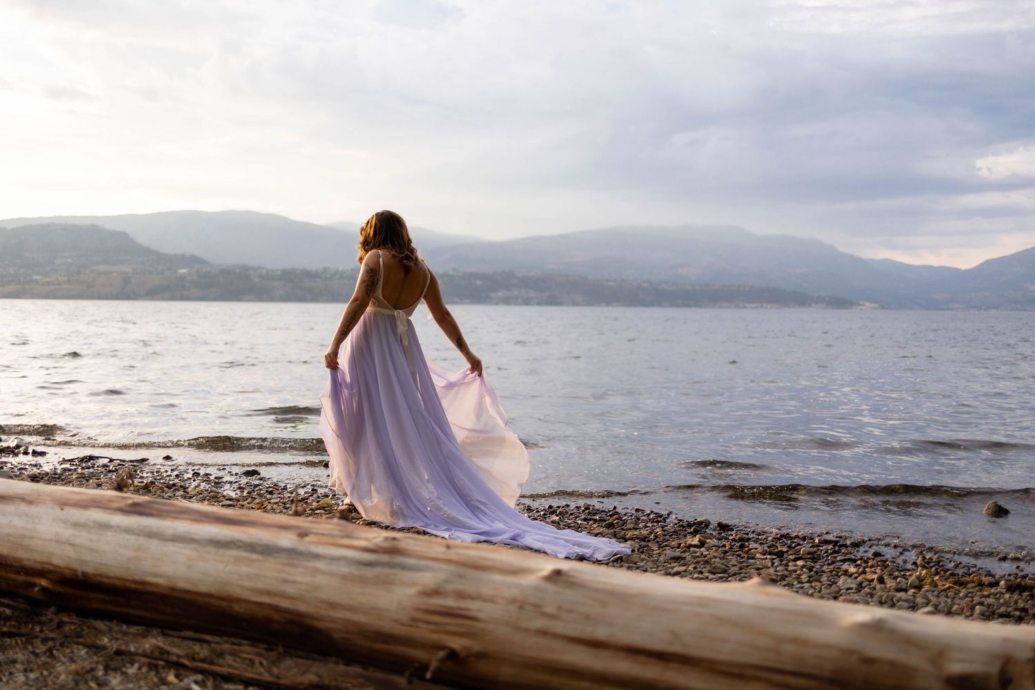 Person in flowing purple dress walking along rocky beach shore with mountain views.