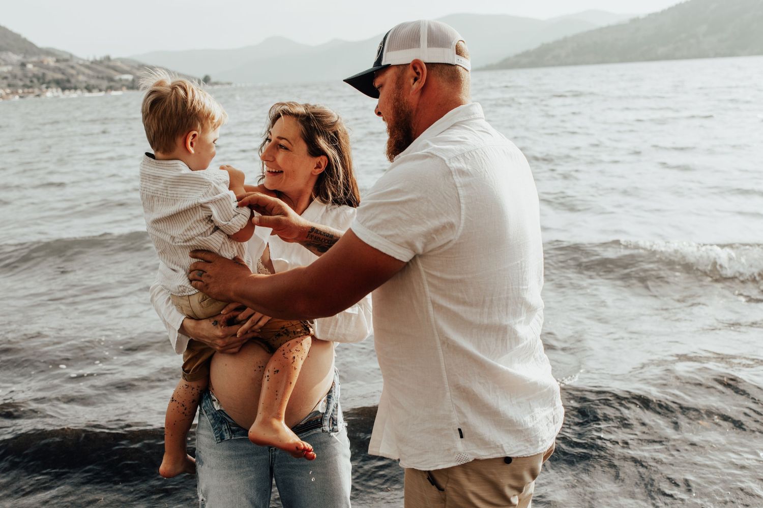 Family enjoying tender moment together on rocky beach with lake and mountains in background.