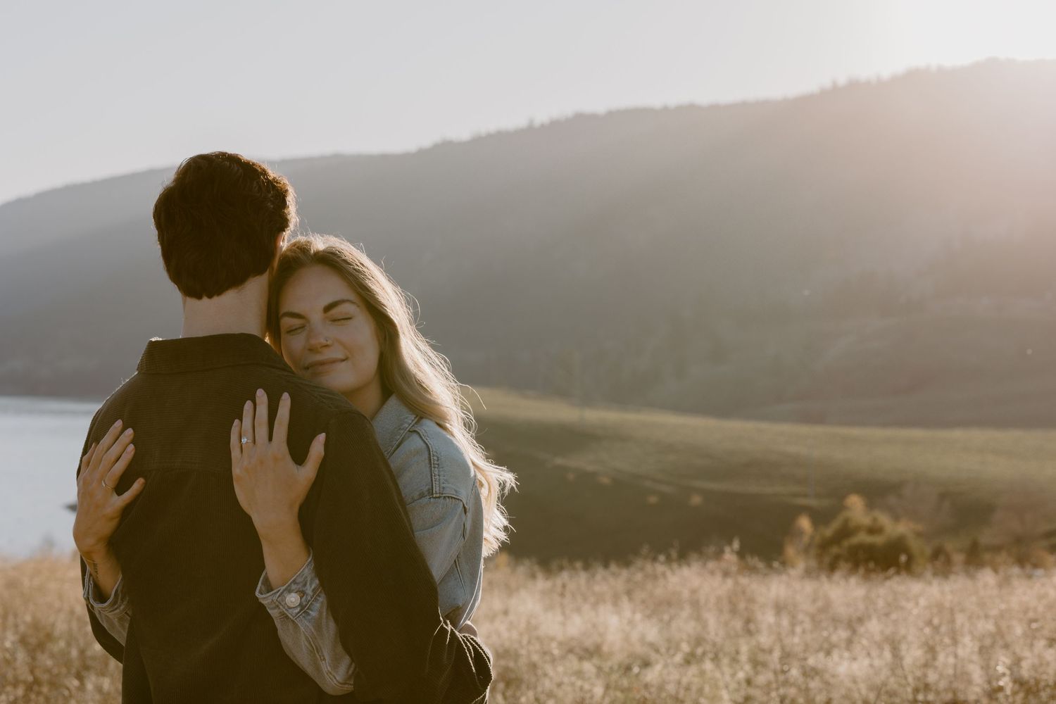 Romantic embrace between couple in golden field with mountains and lake in background.