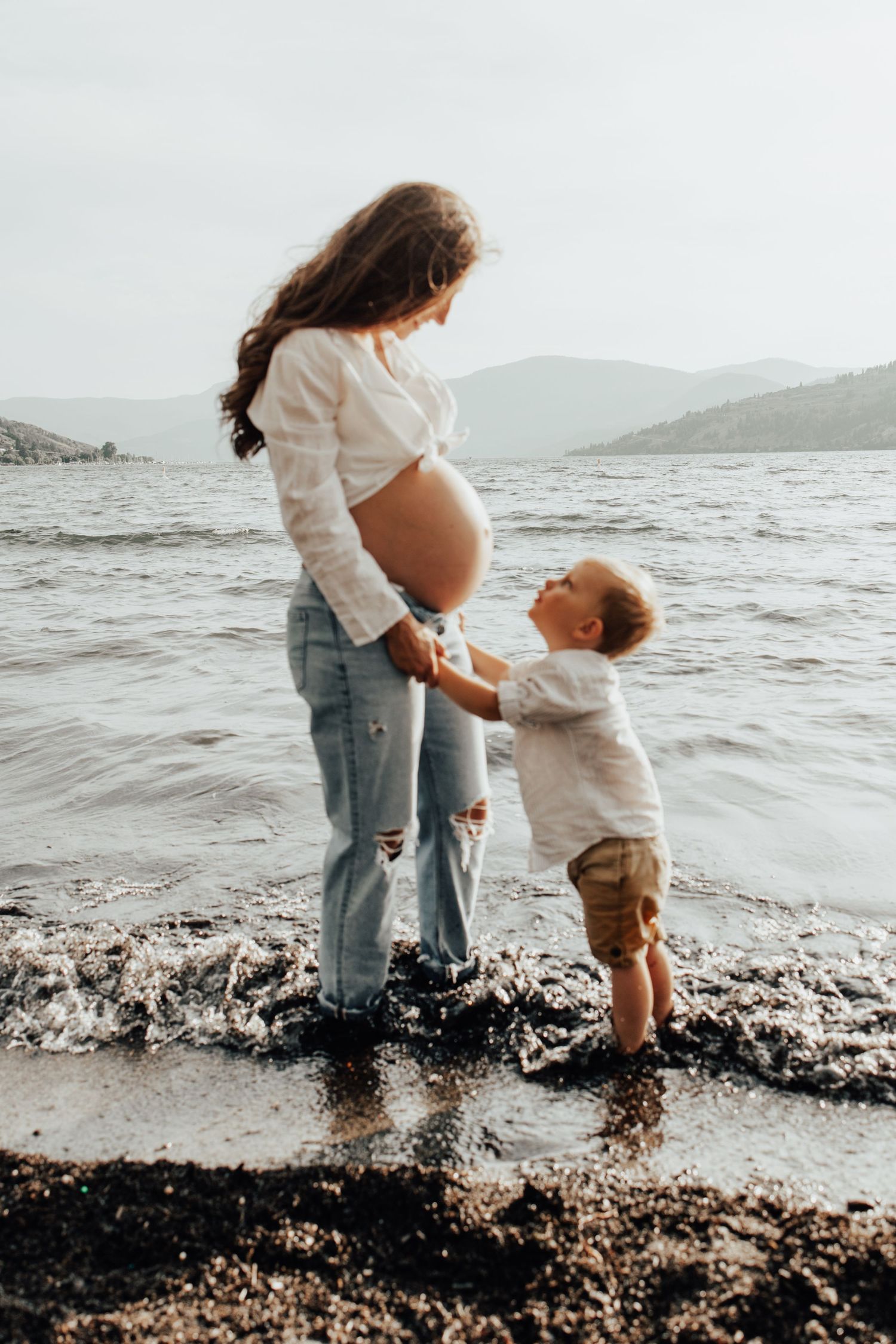 Expectant mother and toddler share moment together at water's edge.