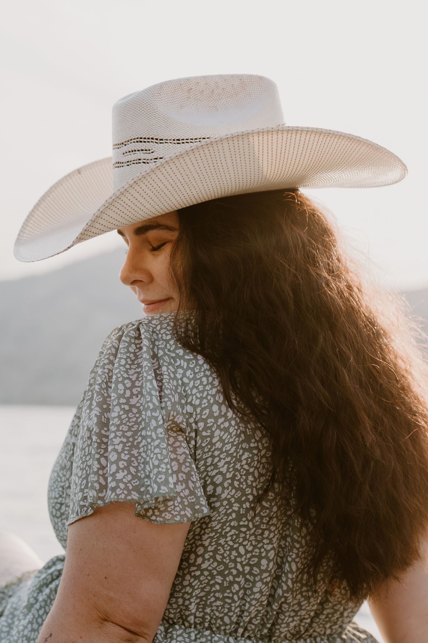 Person in floral dress and white cowboy hat with long dark hair against sunset backdrop.