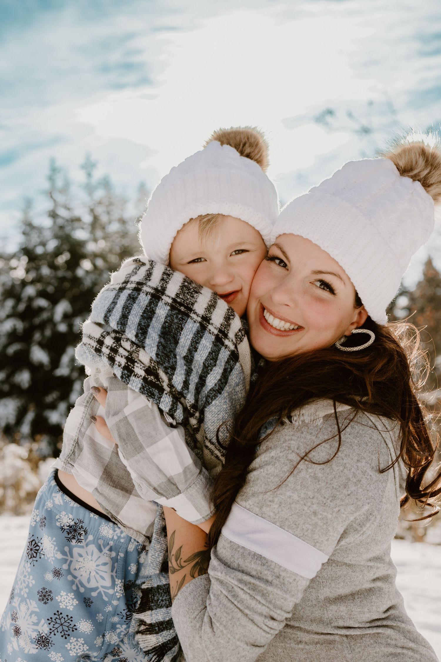 Close-up winter portrait of two people in matching white pom beanies against snowy forest.