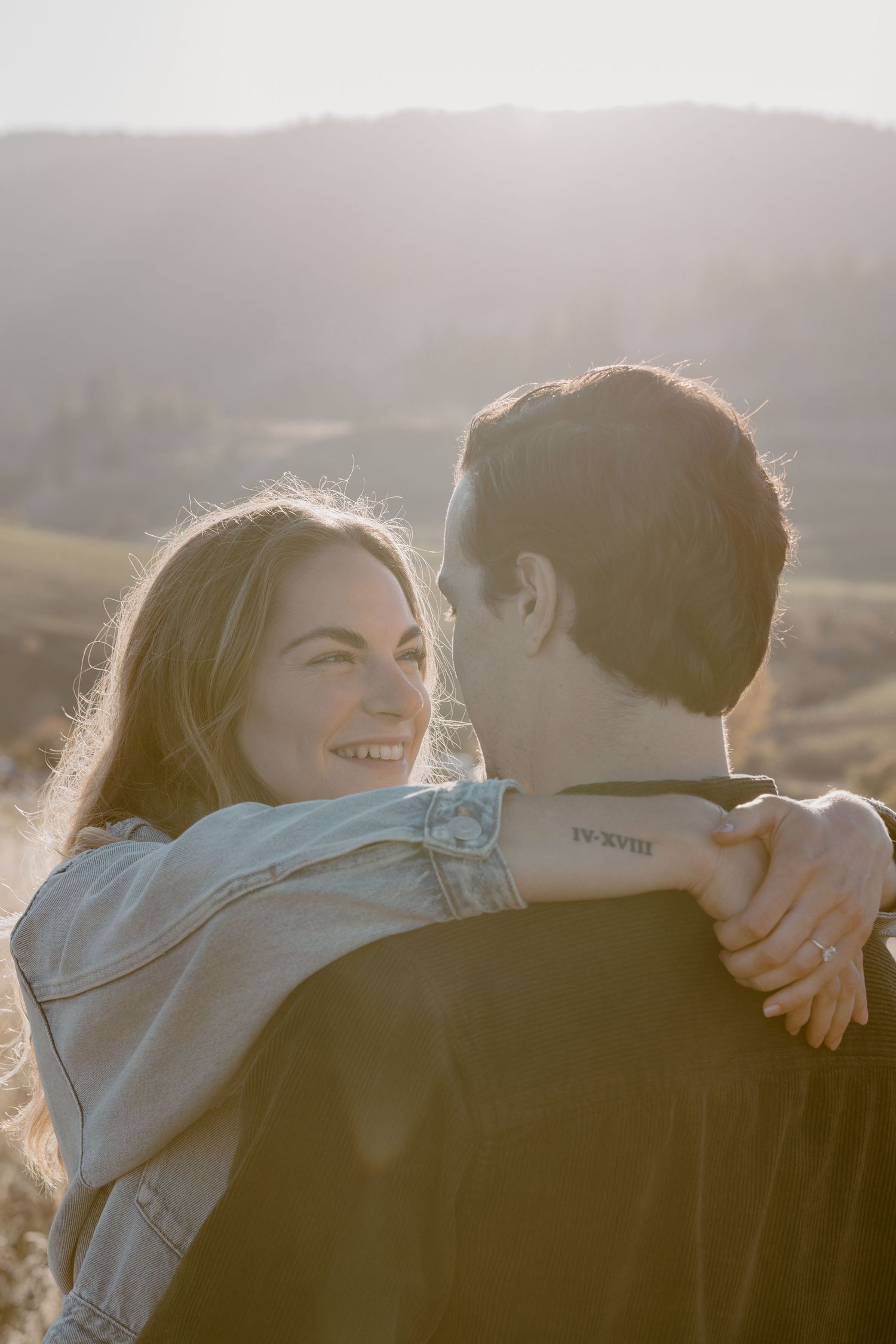 Backlit sunset portrait of couple embracing in casual wear against mountain backdrop.