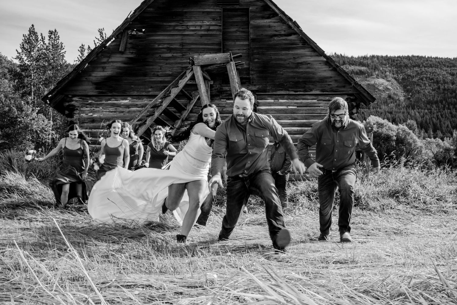 Wedding party running and celebrating playfully in front of rustic wooden barn.