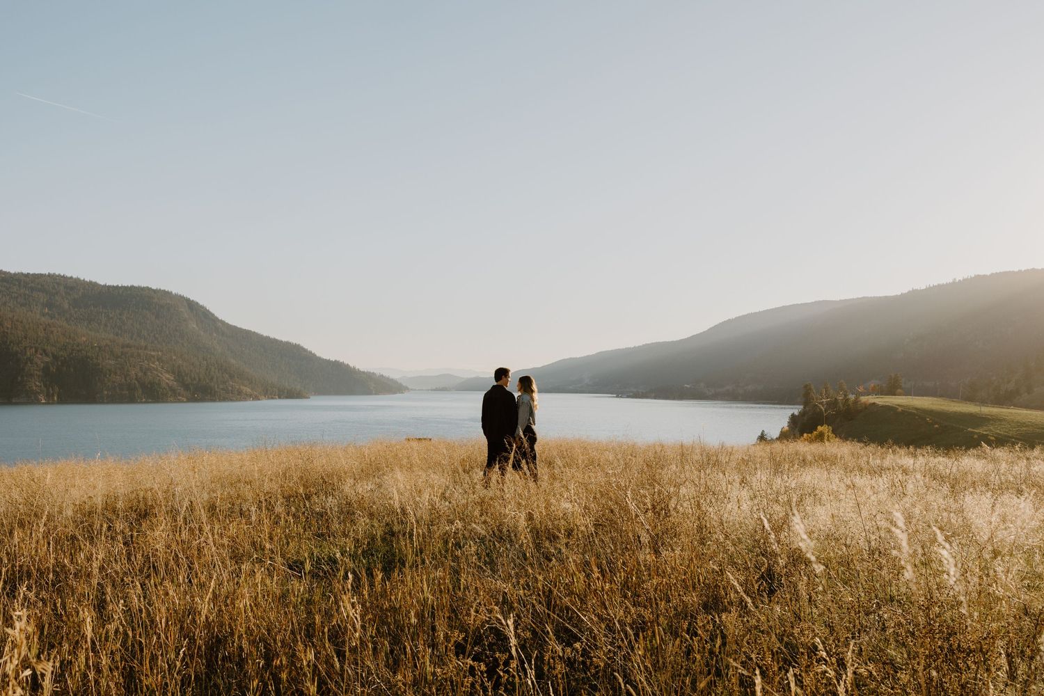 Silhouette of couple standing in tall grass overlooking scenic mountain lake at sunset.