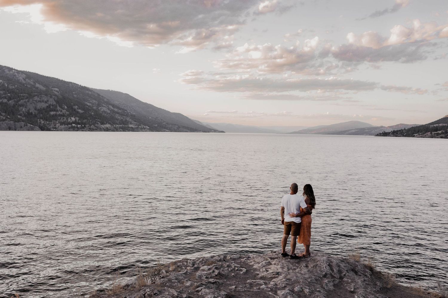 Couple standing on rock overlooking serene lake and mountains at dusk.