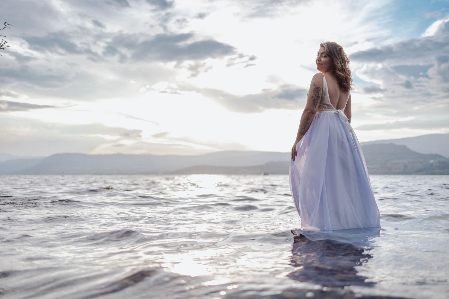 Person in white wedding dress standing in water at sunset with mountains behind.