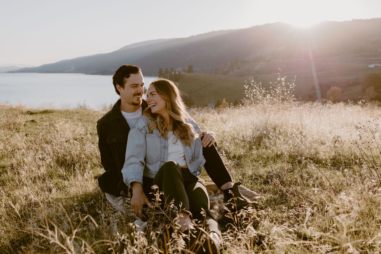 Happy couple sitting together in sunlit meadow with mountains and lake in background.