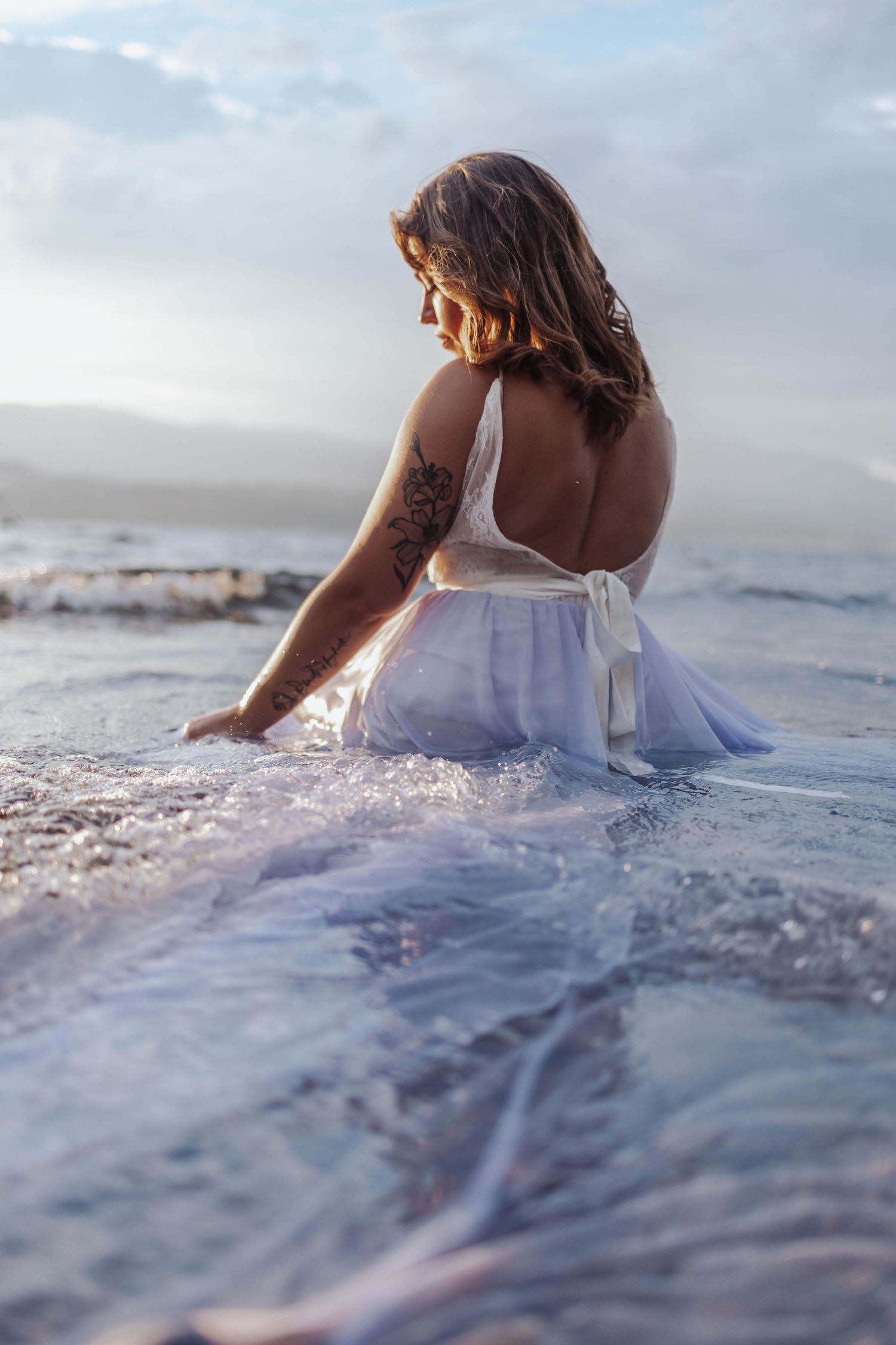 Woman in white dress sitting in ocean waves during sunset creating dreamy coastal atmosphere.